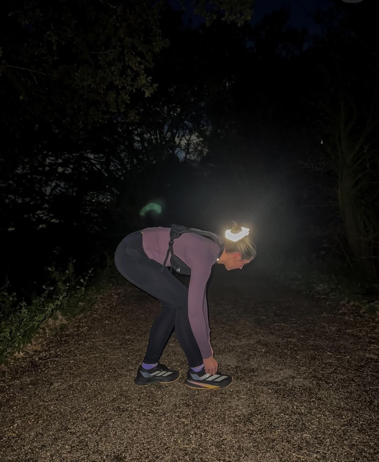 Trail runner crouching to tie a shoe at night, headtorch lit, wearing a reflective Eve Marmalade scrunchie